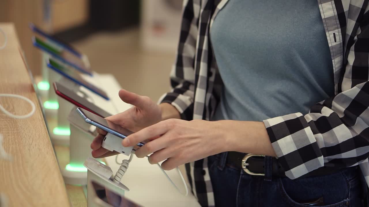 Waist shot of unrecognizable woman's hands chooses a smartphone in an electronics store. She takes smart phone from counter and trying using it. Close up