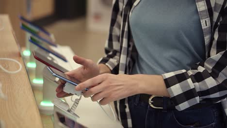 Tiro-De-Cintura-De-Las-Manos-De-Una-Mujer-Irreconocible-Elige-Un-Teléfono-Inteligente-En-Una-Tienda-De-Electrónica.-Ella-Toma-El-Teléfono-Inteligente-Del-Mostrador-E-Intenta-Usarlo.-De-Cerca