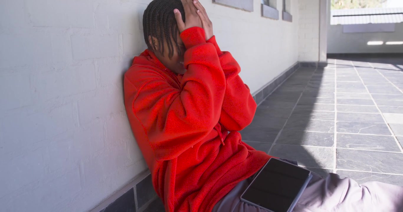 Teen male in red hoodie sitting against brick wall tapping tablet reacting to content, copy space