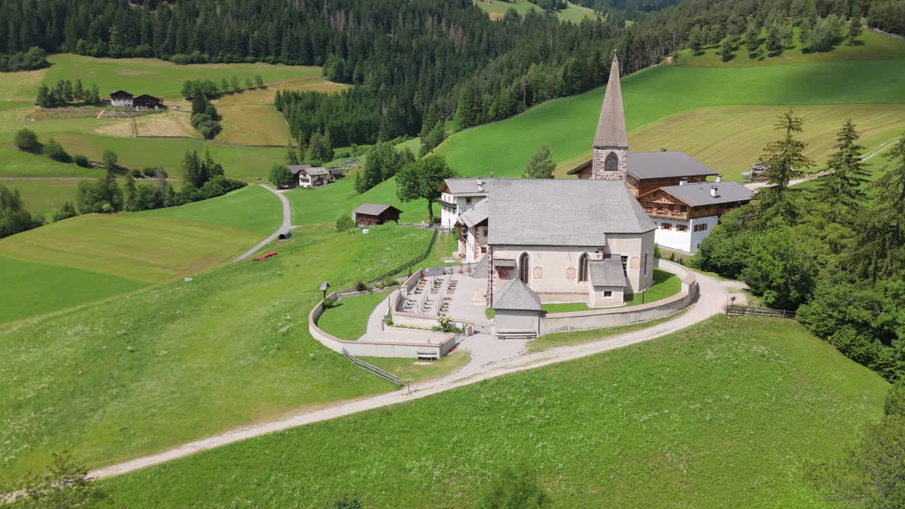 Aerial drone view of a church in the Italian Dolomites, surrounded by green alpine meadows, forested hills and mountain landscape