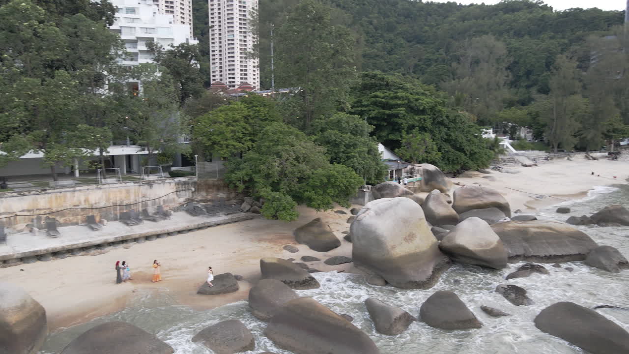 People enjoying Batu Fehrengi beach with restaurant and apartment buildings in the background on the northern side of the island of Penang, Malaysia, panning drone movement.