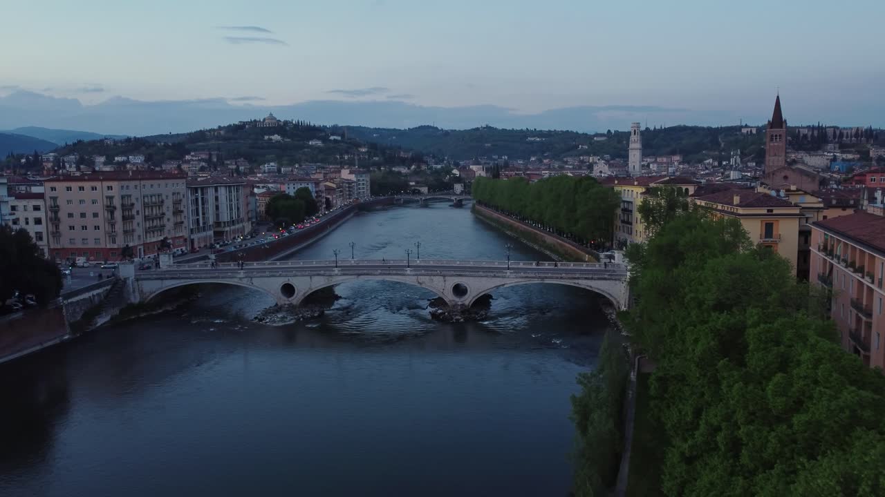 Drone view at blue hour over Ponte della Vittoria spanning the Adige River in Verona, with Sant’Anastasia tower, city lights, and Castel San Pietro hill forming a scenic backdrop
