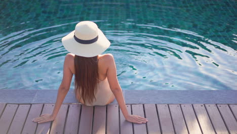 while sitting on the deck of a swimming pool, a young woman, with her back to the camera, kicks her feet in the water