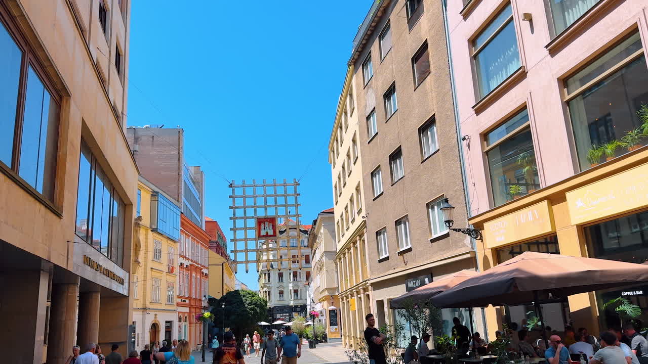 Bratislava, Slovakia - June 5, 2025: Sunny city strolls happening!. People stroll through a vibrant urban street lined with shops and cafes under a clear blue sky