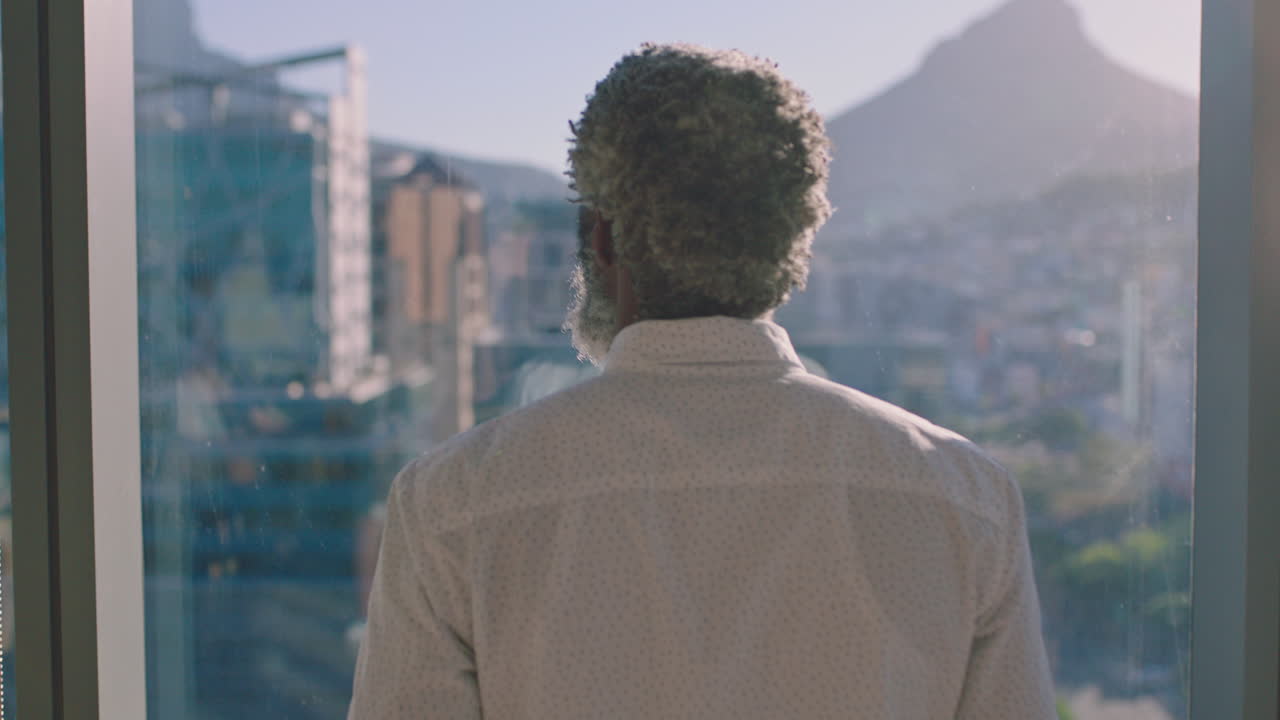 hombre de negocios afroamericano maduro mirando por la ventana planeando hacia adelante pensando en ideas para el desarrollo de negocios futuros con vista a la ciudad al atardecer 4k