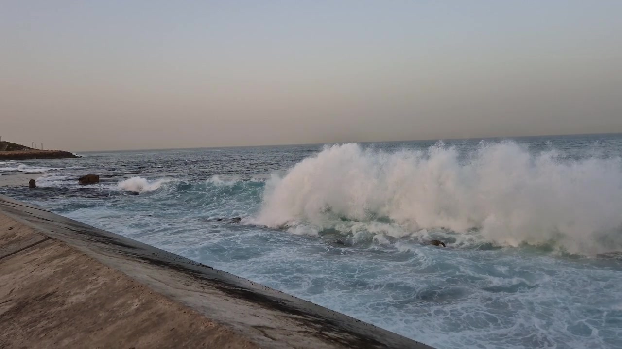 Onde marine spumeggianti si infrangono e schizzano su bassi fondali rocciosi vicino al frangiflutti in cemento dello storico Porto di Giaffa lungo la costa mediterranea sotto un cielo diurno coperto. Panoramica