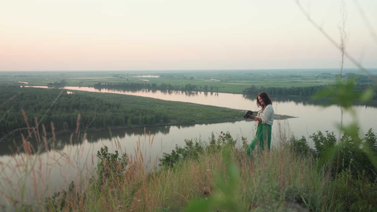 woman photographer checking photo on camera screen while standing on river bank cliff at golden hour with glass cup resting in grass foreground distant river winding through lush green landscape
