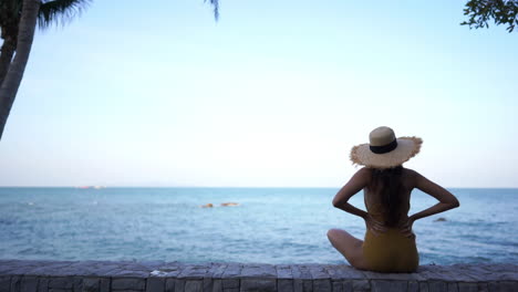 A-young-woman-with-her-hands-on-her-waist-is-sitting-on-a-stone-seawall-looking-out-on-the-ocean-beyond