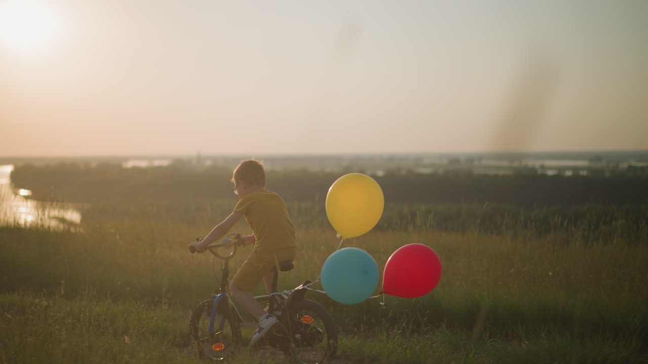 A young boy rides his bicycle through a grassy field at sunset, with vibrant red, blue, and yellow balloons tied to the back of his bike. The scene captures the carefree joy of childhood