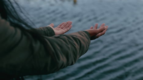 Junge-Frau-Mit-Erhobenen-Armen-Steht-An-Einem-Bewölkten-Tag-Am-See-Und-Fängt-Regen-In-Den-Händen,-Während-Der-Wind-Durch-Ihre-Haare-Weht