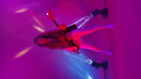 Vertical-video-of-a-dancing-cheerful-woman-playing-a-guitar-in-the-changing-background-light-and-strobe-lights-of-a-concert-light