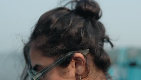 primer plano de una mujer joven en la playa, mano en el cabello, con gafas de sol que reflejan el cielo azul, estado de ánimo contemplativo