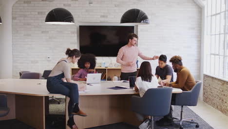 Millennial-white-man-stands-addressing-his-creative-colleagues-in-a-meeting-room,-full-length