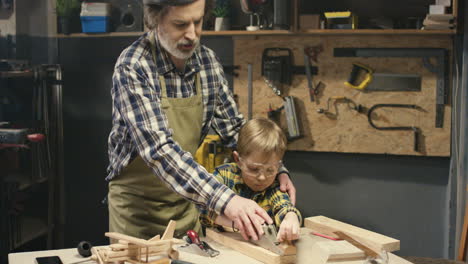 viejo carpintero caucásico con barba gris enseñando a su nieto a trabajar con madera dura en el taller