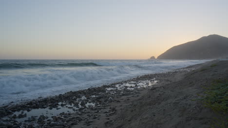 Toma-Estacionaria-De-Olas-Rompiendo-Contra-La-Costa-Rocosa-De-La-Playa-De-Mondo-Con-Puesta-De-Sol-Detrás-De-Una-Montaña-Ubicada-En-El-Sur-De-California