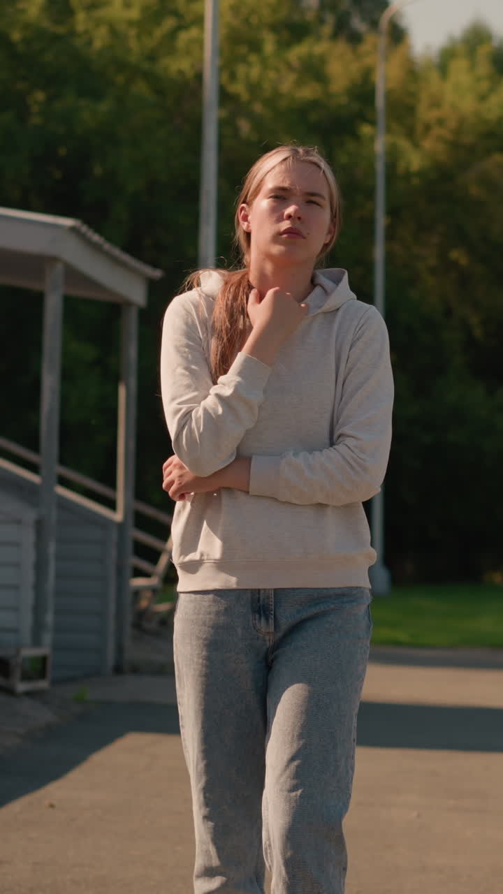 una joven con una sudadera casual camina pensativa cerca de un estadio vacío con la mano en su sudadera, rodeada de árboles verdes y postes eléctricos