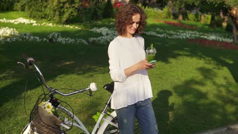 portrait of a young woman surfing in the internet using her smartphone standing in the city park near her city bicycle with