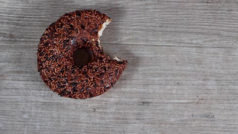 a delicious chocolate doughnut with chocolate sprinkles having a bite taken out of it and put back down on a wooden work top