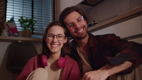 Portrait-of-a-Happy-duo-young-brunette-girl-in-glasses-and-wireless-red-headphones-together-with-her-brunette-boyfriend-with-stubble-sitting-on-the-floor-in-a-modern-apartment-in-the-kitchen-and-posing