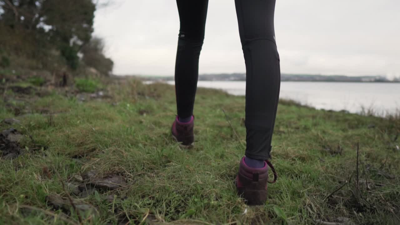 Slow motion close up shot from the back of girl walking on the grassy and rocky ground on the coast of a lake