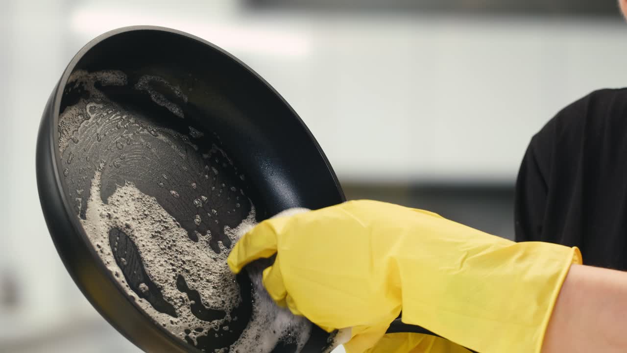 Nahaufnahme einer Hand mit gelbem Handschuh, die eine Bratpfanne spült