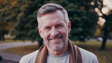 mature man wearing casual clothes and brown scarf looking at camera and smiling, expressing positive emotions such as happiness, joy and optimism, in a park during a sunny autumn day