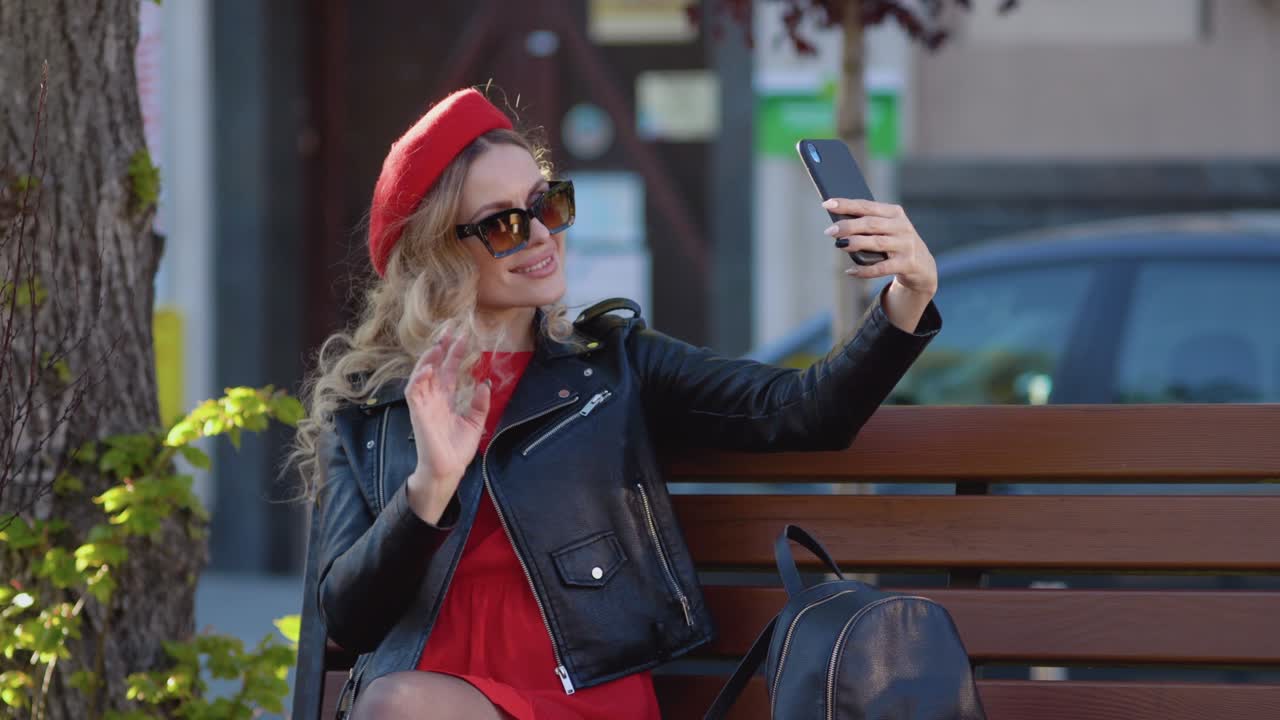Young woman in stylish clothes taking a selfie sitting on a park bench. Smartphone as a universal gadget