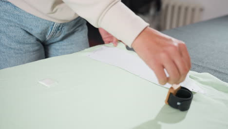 woman in jeans carefully joins two fabrics using office pin on workspace table, securing white paper pattern over light green textile during professional garment preparation process