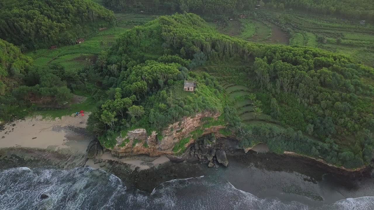 aerial view of a coastal cliff covered with a forest and on top a house looking down on a beach where the waves break on the sand