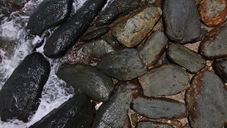 toma estática aérea de rocas oscuras en una playa con pequeñas olas rompiendo suavemente sobre ellas