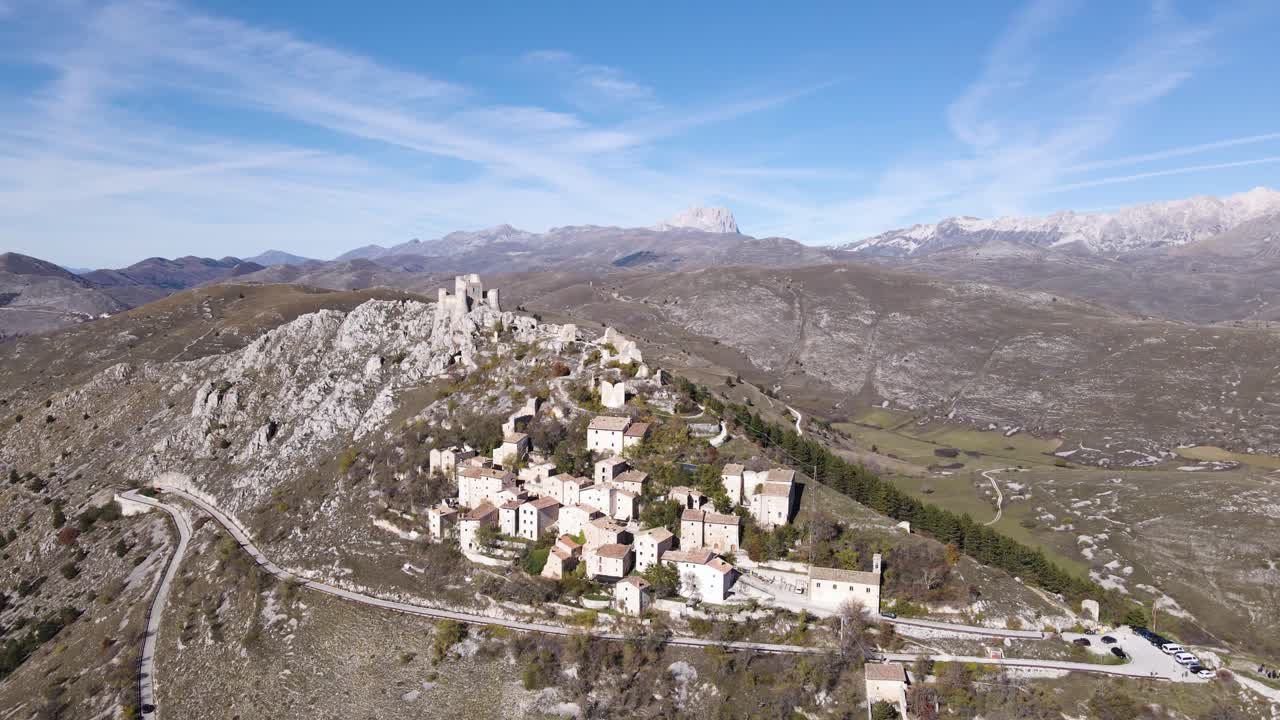 Splendida vista aerea della fortezza medievale di Rocca Calascio e del borgo arroccato su uno sperone roccioso, con la catena montuosa innevata del Gran Sasso visibile nel paesaggio abruzzese.