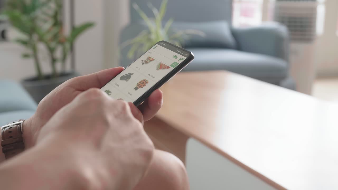 Man Sitting On Couch Using Smartphone Buys In Internet Shop. Male Selects Vegetables In Grocery Online Store