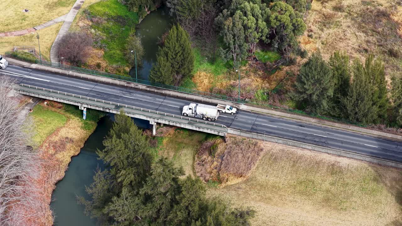 Overhead drone footage captures multiple semi-trucks and cars traveling across a rural bridge above a river, with natural daylight and steady camera movement