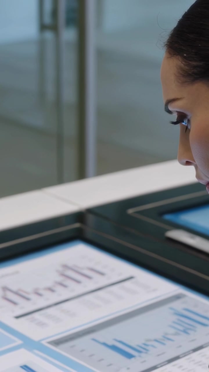 Vertical video: Entering right, woman in suit leaning over touch table studying charts, copy space