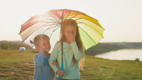 boy and girl protected by rainbow parasol siblings unhappy because of drops on face standing beneath