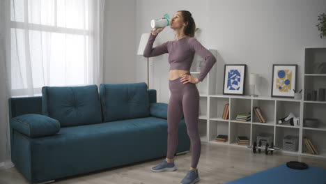 Una-Mujer-Deportista-Está-Parada-En-La-Sala-De-Estar-Y-Descansando-Con-Agua-Potable-En-El-Entrenamiento-Matutino-En-Casa,-Fitness-Y-Bienestar.