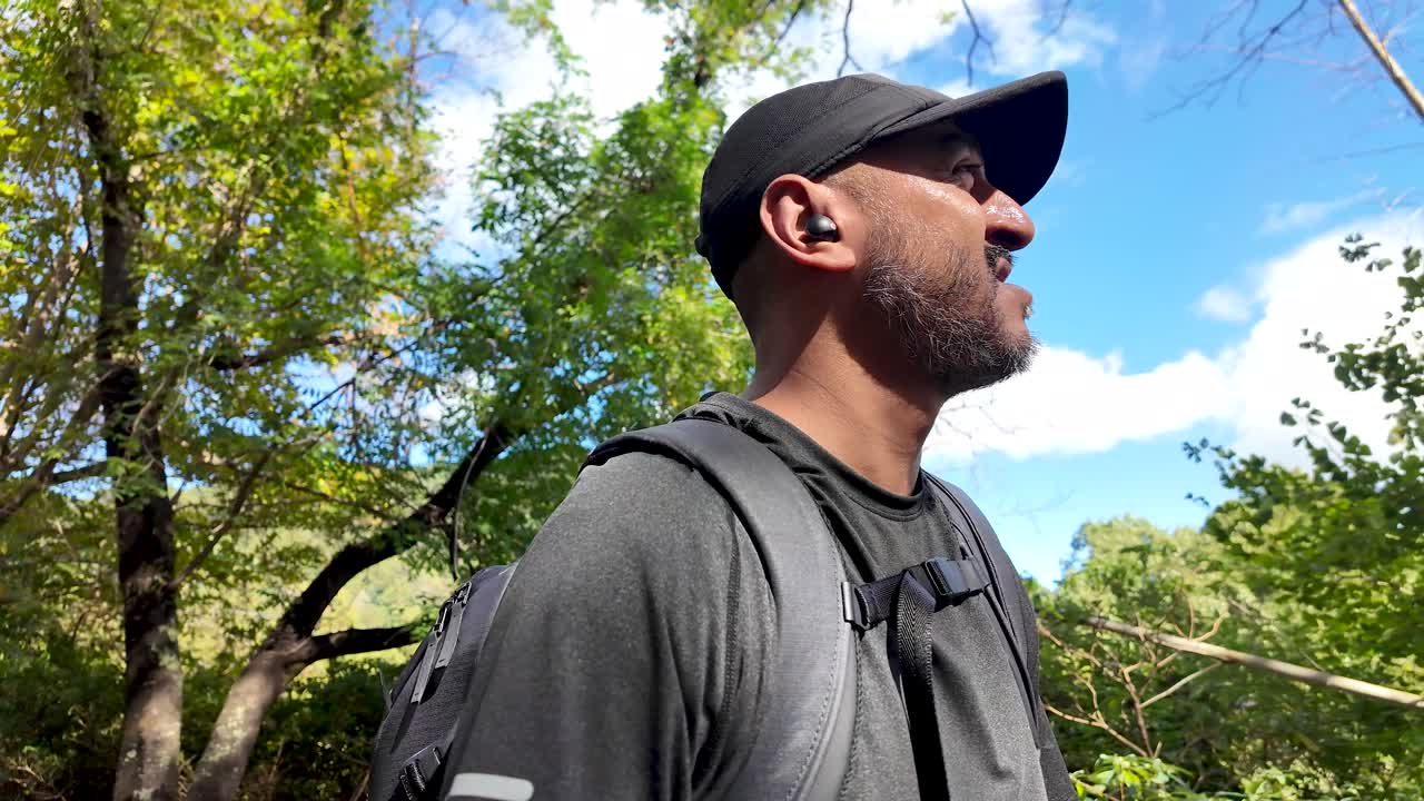 South asian hiker with backpack and wireless earbuds enjoying trekking in forest during sunny day. slow motion