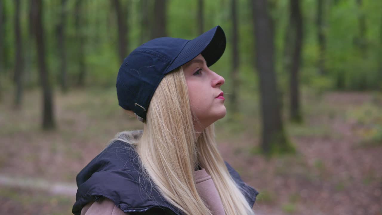 A young blonde woman wearing a dark cap and casual outdoor clothing stands in a forest, looking thoughtfully to the side. The natural green background creates a calm and peaceful atmosphere