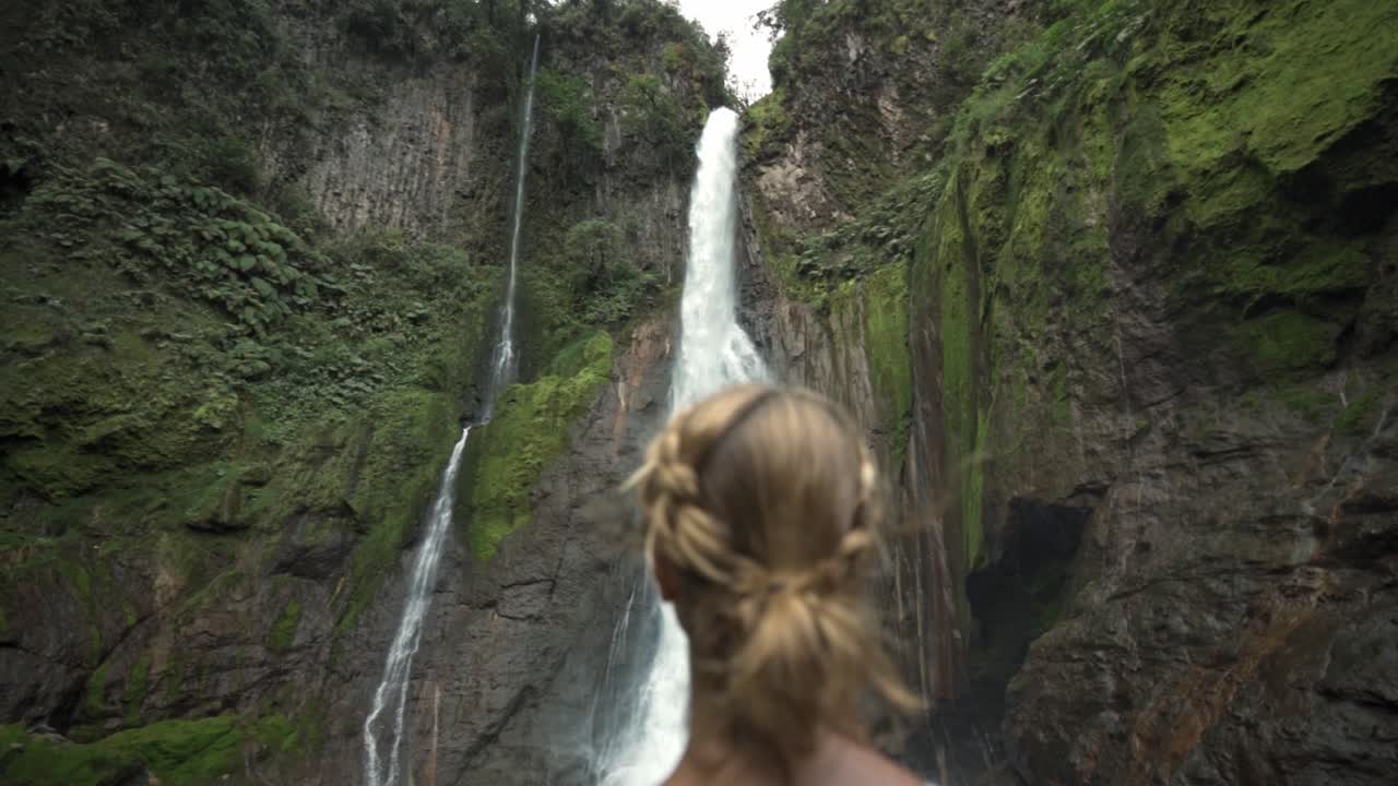 A woman, seen from behind, standing in a lush rainforest and looking out at the majestic Catarata del Toro waterfall in Costa Rica