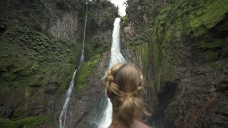 a woman, seen from behind, standing in a lush rainforest and looking out at the majestic catarata del toro waterfall in costa rica