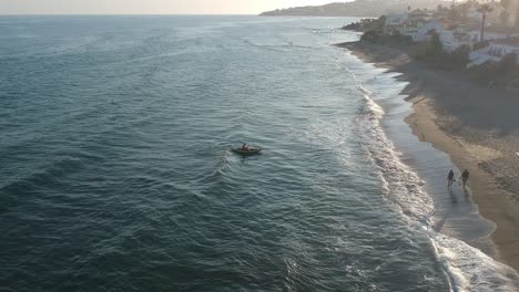 Vistas-Aéreas-De-Una-Playa-Con-Un-Hombre-En-Un-Kayak-Y-Gente-Caminando-Por-La-Orilla