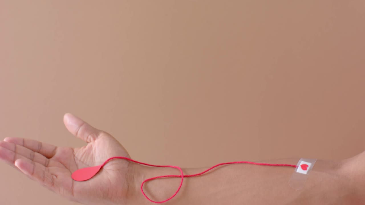 mano de hombre biracial con gota de sangre, cuerda roja y corazón pegando yeso en beige, cámara lenta