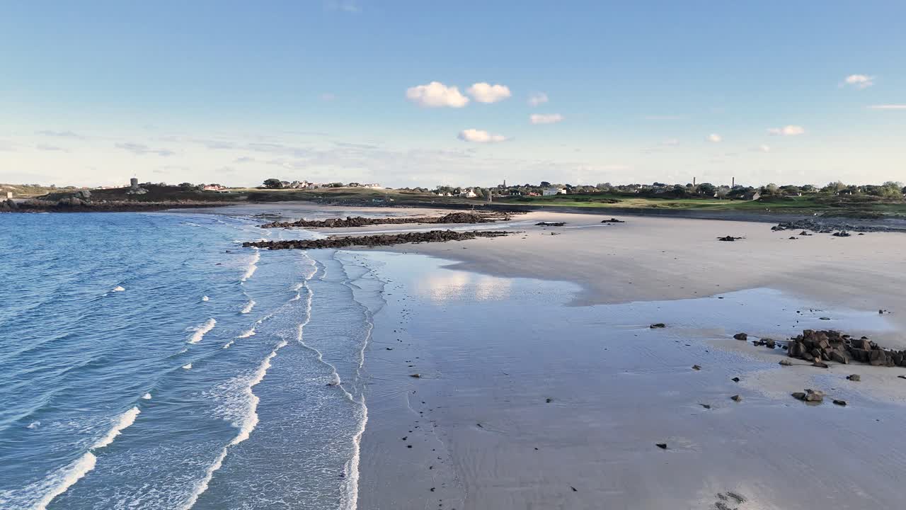 Lancresse Bay Guernsey low forward flight over shoreline of golden sandy beach with rocky outcrops in late afternoon sun with calm lapping waves and views over Lancresse Common