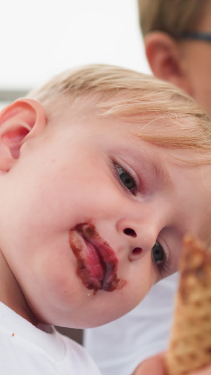 Two young brothers are sitting together, savoring their ice cream cones, the younger one, with chocolate smeared on his mouth, intently enjoys his cone while the older one takes a big bite