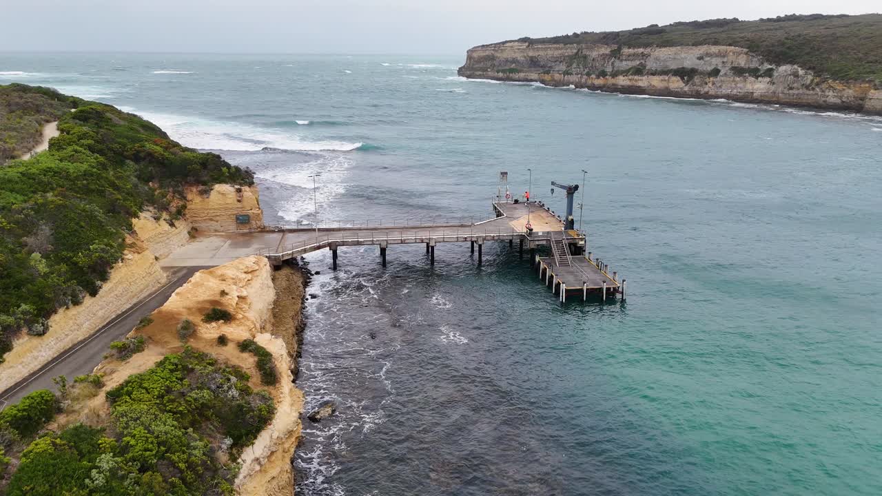 Drone footage captures Port Campbell's pier and rugged coastline. Overcast skies and calm waters create a serene, natural atmosphere
