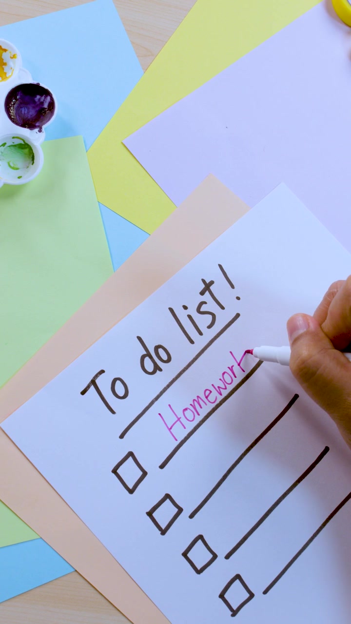 Person skriver 'Lektier' på en to-do-liste, omgivet af farverigt papir, blyant og malerpalet