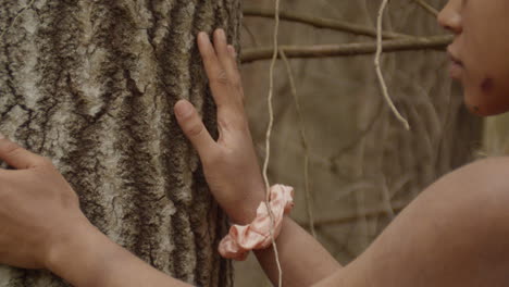 The hands of an ebony girl run along the bark of a sweetgum tree. A pink scrunchie on her right wrist. Right hand runs down along the bark, while left hand goes sideways, away from her. .
