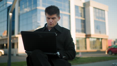 focused investor seated outdoors operating laptop with leg crossed, background features modern glass building, parked cars, and greenery