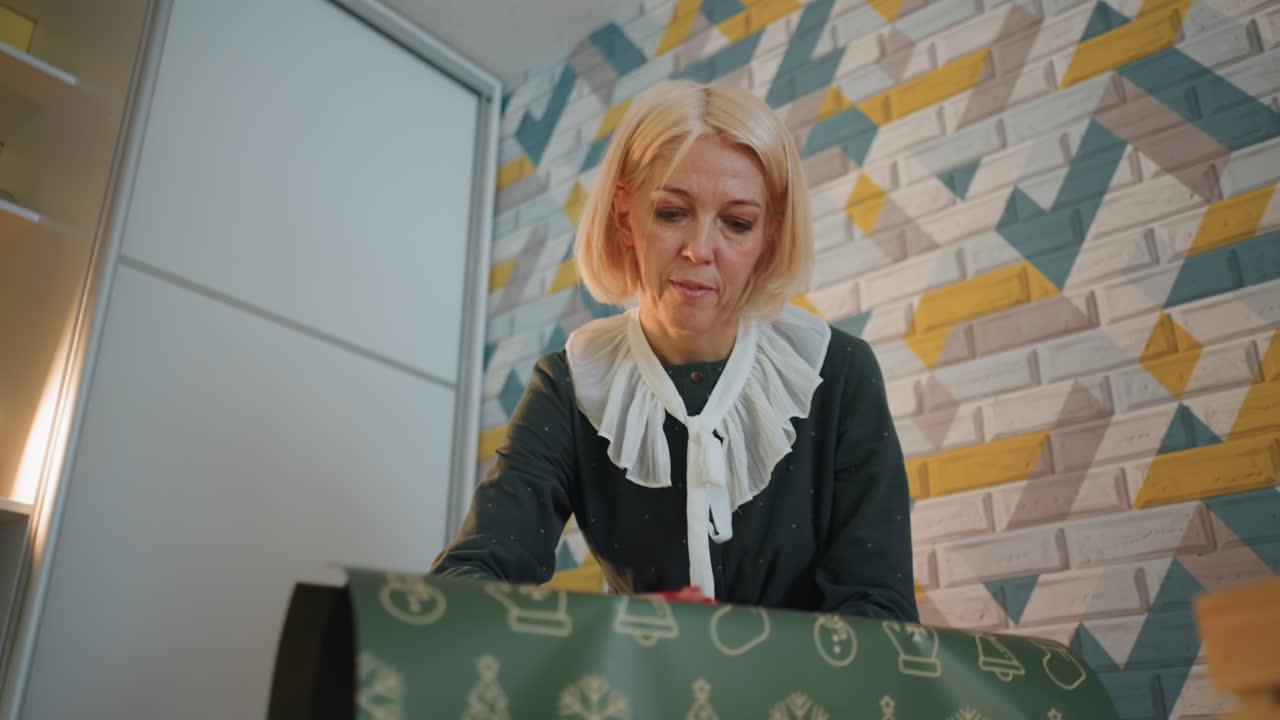 Frau verpackt Weihnachtsgeschenk auf dem Tisch, konzentrierter Ausdruck, gemusterte Wand als Hintergrund, weiches warmes Licht, Band und Schere, festliche Bastelszene, aufgeräumter Schreibtisch, Nahaufnahme der Geschenkvorbereitung.