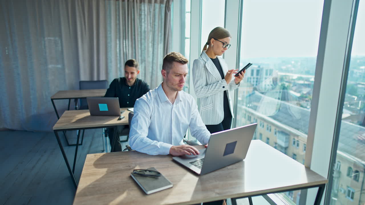 Serious focused man in white shirt working at laptop. Female colleague looks at her phone standing at the window. Office indoors with panoramic window at backdrop.